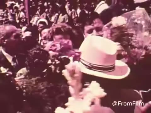 The still from the 'Project Hope Public Service Announcement' features a crowd scene, indicative of the 60s/70s era, with people engaging closely and displaying various emotions. In the foreground, a person wearing a white hat is prominently visible, surrounded by individuals holding flowers. The atmosphere is vibrant, suggesting a moment of celebration or gratitude related to health initiatives. This visual encapsulates Project HOPE's mission to provide health care globally, highlighting human connection and community support during that time.