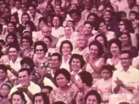 The still from the 'Project Hope Public Service Announcement - 60's/70's' features a diverse crowd of people, primarily smiling and applauding. The audience includes men and women of various ages, dressed in colorful attire characteristic of the time. The setting appears to be an event or gathering, possibly related to health initiatives, reflecting the spirit of community engagement that Project HOPE embodied through its mission of providing health care worldwide.