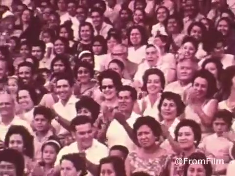 The still from the 'Project Hope Public Service Announcement' features a large crowd of people, representing a diverse audience from the 1960s or 70s, gathered in what appears to be a public event or gathering. Many individuals are smiling and clapping, showcasing a sense of community and engagement. This imagery aligns with the mission of Project HOPE, highlighting its focus on health and humanitarian efforts. The overall atmosphere conveys enthusiasm and support for the cause.