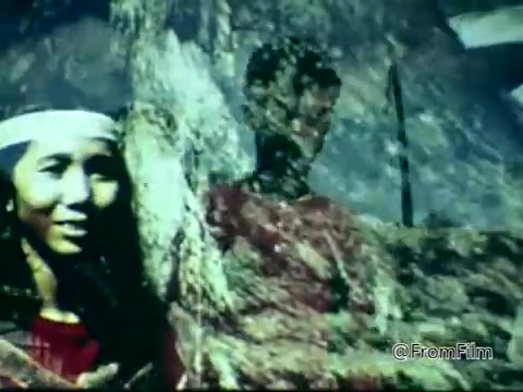 The still from the 1975 PSA by the Columbian Fathers features a smiling woman in traditional attire, set against a blurred background that suggests a natural or cultural landscape. The image appears vibrant and layered, possibly employing a composite effect to convey a message related to the mission work or cultural themes pertinent to the organization's outreach efforts in Colombia.