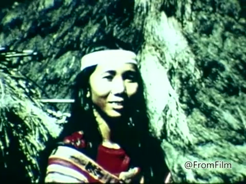 The still from 'Columbian Fathers PSA - 1975' features a young woman with long hair and a headband, standing near a rock formation. She is dressed in traditional attire characterized by vibrant colors and patterns, suggesting an indigenous cultural background. The image conveys a sense of connection to nature, potentially highlighting themes of heritage and identity relevant to the PSA's message.