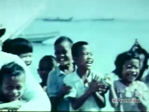 The still from the 'Columbian Fathers PSA - 1975' features a group of children laughing and smiling. They appear to be outdoors, with a body of water and boats in the background. The scene conveys a sense of joy and community, reflecting the positive impact of the Columbian Fathers' outreach efforts. The colors in the image are muted, typical of film from that era.