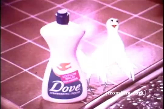 The still features a bottle of Dove Dishwashing Liquid prominently displayed on a tiled kitchen countertop. Next to the bottle is a small animated dove, adding a whimsical touch to the advertisement. The overall color palette appears warm and nostalgic, reflecting the 1970s aesthetic. The product branding is clear, emphasizing its role as an effective cleaning solution.