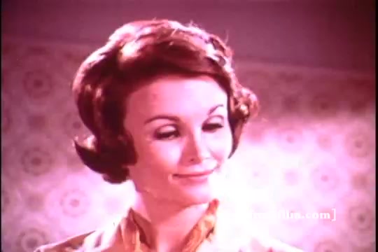 The still features a woman with a stylish 1970s hairstyle, smiling knowingly with a hint of charm. She appears in a bright, soft-lit interior, characterized by a vintage floral-patterned wallpaper that evokes a domestic setting. Her attire reflects the fashion of the era, emphasizing a warm and welcoming ambiance typical of advertising from that time. The overall tone suggests a light-hearted appeal to homemakers, promoting the benefits of Dove Dishwashing liquid.