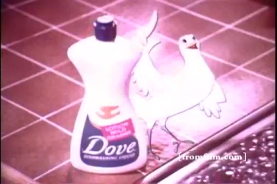 The still features a bottle of Dove dishwashing liquid prominently placed against a tiled kitchen background. Next to it is a cartoonish, animated white dove, playfully depicted, enhancing the product's branding. The scene captures a lighthearted, inviting atmosphere typical of 1970s advertising, emphasizing cleanliness and gentle care.
