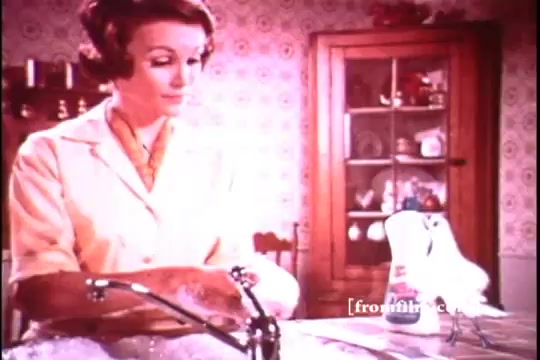 The still depicts a woman in a vintage kitchen, presumably demonstrating Dove Dishwashing Liquid from the 1970s. She is focused on washing dishes, surrounded by bubbles, while a bottle of Dove sits prominently on the counter. The kitchen features retro wallpaper and a wooden cupboard displaying dishes, highlighting the domestic and cheerful atmosphere typical of that era's advertising. The overall tone is warm, emphasizing cleanliness and ease of use.