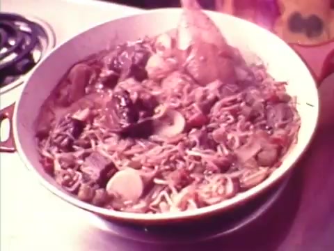 The still features a vibrant dish of noodles and mixed vegetables in a large, round pan. The ingredients appear steaming and include pieces of meat and various colorful vegetables, showcasing a hearty meal. The kitchen background suggests a homey setting, typical of food commercials from the 1970s, highlighting a warm, inviting atmosphere.