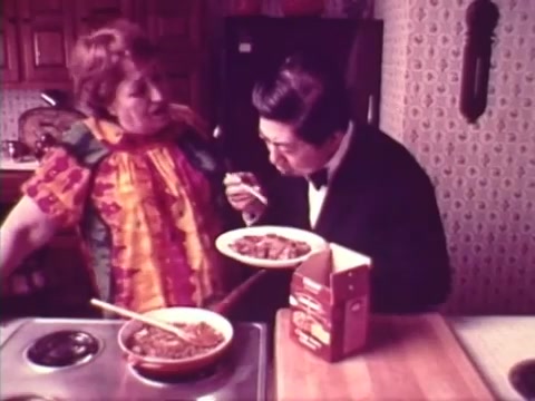 The still captures a scene from the 1973 'Chun King' Chinese food commercial featuring a woman, likely Mama Cass's mother, playfully interacting with Charles Louie. She wears a colorful patterned blouse while Louie, dressed in formal attire, holds a plate of food, seemingly enjoying a taste test. The kitchen setting is cozy, with wooden cabinets and a patterned wallpaper, emphasizing a warm, familial atmosphere.