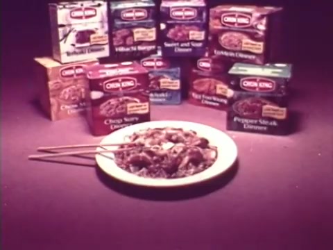 The still features a plate of food with chopsticks in front, showcasing a dish likely from a Chun King Chinese food commercial. Behind it are various colorful product boxes of Chun King meals, such as 'Chop Suey Dinner' and 'Pepper Steak Dinner.' The overall presentation highlights the convenience of these frozen meals, emphasizing their appeal for easy home dining. The nostalgic aesthetic reflects the era of the 1970s.