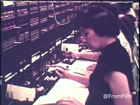 The still from the 16mm print depicts a telephone operator at a busy switchboard in the 1970s, engaged in explaining long-distance calling rates. She focuses intently on her task while surrounded by a complex network of wires and connections. The scene captures the vintage aesthetic and emphasizes the operator's role in facilitating communication, particularly highlighting the advice to make calls before the rates increase on Sunday evening. The overall atmosphere conveys a sense of urgency and dedication typical of telephone operators during that era.