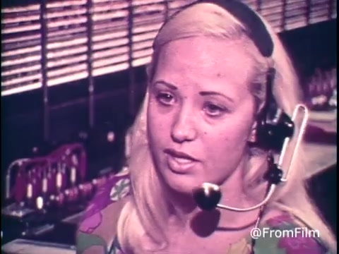 The still captures a telephone operator in a period-appropriate work environment, wearing a headset with a microphone. She has styled blonde hair and is dressed in a colorful floral outfit typical of the 1970s. The backdrop features a row of telephone switchboards, indicative of the technology of the time. The operator appears engaged as she explains to viewers the nuances of long-distance calling rates, specifically advising them to make calls on Saturday or prior to 5 PM on Sunday to avoid higher charges. The overall tone portrays a sense of helpfulness and customer service.