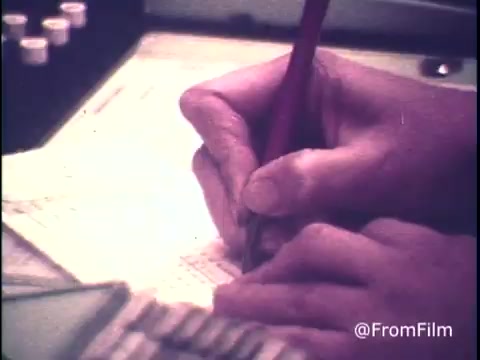 The still captures a close-up of a telephone operator's hands as they jot down notes or information, likely regarding long-distance call rates. The setting suggests a busy telecommunications environment typical of the 1970s, with equipment and documentation visible in the background. The tone of the scene reflects a focused and informative atmosphere, emphasizing the operator's role in guiding customers about cost-effective calling times.