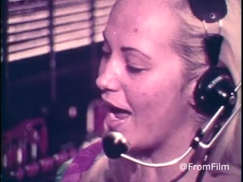 The still features a telephone operator with light blonde hair and a headset, speaking directly to the camera. She appears engaged as she explains long-distance calling rates. In the background, a traditional telephone switchboard can be seen, emphasizing the vintage context of the 1970s. The operator advises callers to make their long-distance calls on Saturday and throughout Sunday until 5 PM to avoid higher rates, highlighting the nuances of cost management during that era. The color and quality of the 16mm print reflect the period's aesthetics.