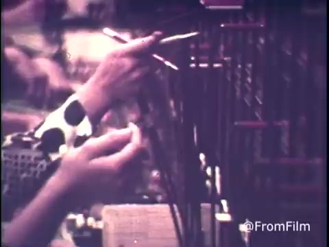 The still from 'C&P Telephone - Operator talks Long Distance Rates - 1970's' features a close-up of a telephone operator's hands maneuvering through a complex switchboard system. The operator is likely explaining the nuances of long-distance calling rates, emphasizing that rates increase on Sundays at 5 PM. In her patterned blouse, she uses a mix of traditional equipment to manage incoming and outgoing calls, reflecting the vintage telecommunication technology of the era. The image captures the busy and service-oriented atmosphere of a telephone exchange during that time.