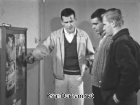 The still depicts three young men engaged in conversation while looking at a recruitment poster for the US Marine Corps. One man, dressed in a light-colored jacket, points at the poster, indicating a discussion about military roles. The other two men appear attentive, dressed in casual sweaters. The background suggests a simple indoor setting, likely a school or community space, emphasizing the focus on recruiting youth during the Vietnam War era.