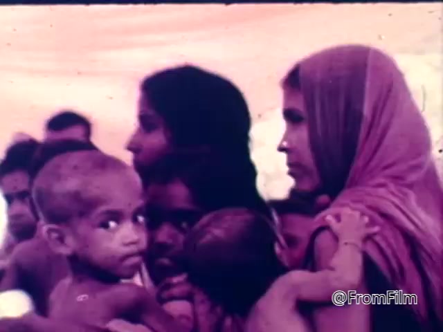 The still depicts a somber scene from the 1975 CARE World Hunger Fund PSA, showcasing a group of vulnerable individuals, primarily women and children. The expressions on their faces convey distress and concern, highlighting the impact of hunger and poverty. The use of colors suggests a muted, serious tone, emphasizing the urgency of the issue being addressed. Overall, the image captures the emotional weight of a humanitarian crisis.