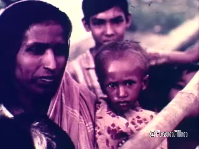 The still from the 1975 CARE World Hunger Fund PSA features a close-up of a woman and a child, with another boy slightly behind them. The woman's expression appears serious, reflecting concern, while the child gazes directly at the camera with a serious demeanor. The background suggests rural or impoverished surroundings, emphasizing the theme of hunger and need for aid in the PSA. The overall tone conveys urgency and a call for compassion towards those affected by hunger.
