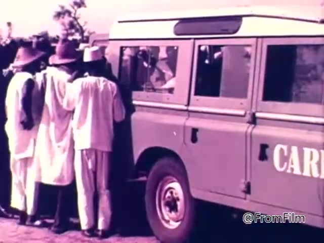 The still from the 1975 CARE World Hunger Fund PSA depicts a group of individuals, dressed in traditional attire, gathered around a vehicle marked with 'CARE.' The scene suggests a moment of interaction, likely involving aid distribution or support related to hunger issues. The warm color palette conveys a sense of urgency and humanitarian effort.
