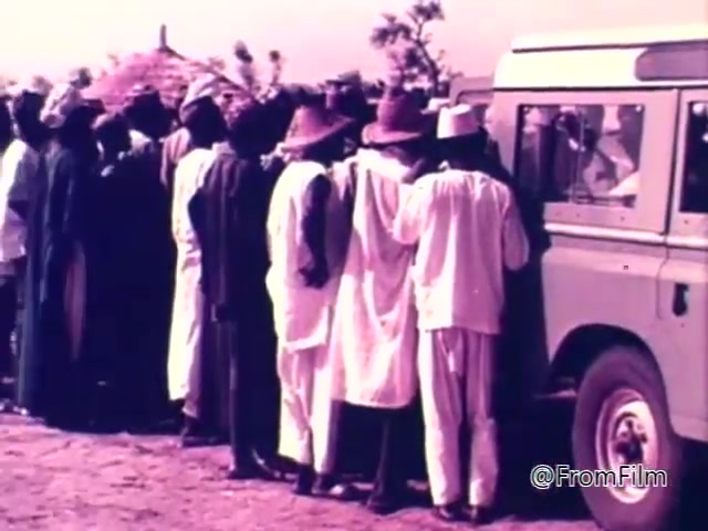 The still from the 1975 CARE World Hunger Fund PSA depicts a group of people, primarily men, gathered around a vehicle, likely waiting for aid or supplies. They are dressed in traditional clothing, with some wearing wide-brimmed hats, suggesting they are in a rural setting. The background features sparse vegetation and huts, highlighting the context of the humanitarian effort focused on addressing hunger. The overall tone conveys urgency and community need.