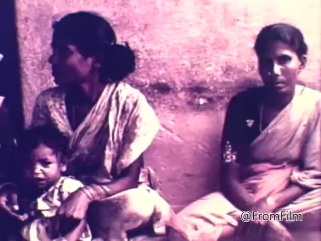The still depicts two women seated against a wall, with one woman holding a child. Both women are dressed in traditional attire, conveying a sense of cultural context. Their expressions suggest concern and resilience, reflecting the themes of hardship and the impact of hunger that the PSA aims to address. The color tone is muted, indicative of the era, emphasizing the seriousness of the message related to world hunger.