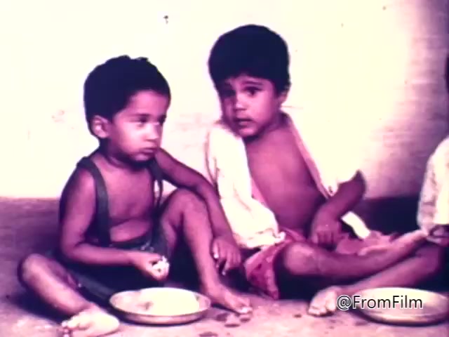 The still from the 1975 CARE World Hunger Fund PSA depicts two young children sitting on the ground in a bare environment. One child, dressed in a green tank top, holds food while looking thoughtfully, and the other, wearing a loose white garment, gazes at him. A small, empty plate is placed between them, highlighting themes of hunger and poverty. The image evokes emotional responses related to childhood vulnerability and the need for humanitarian aid.