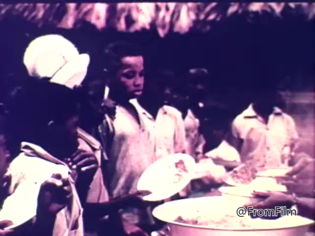The still from the 1975 CARE World Hunger Fund PSA depicts a group of children, primarily boys, gathering around a food distribution point. They are waiting to receive meals, holding empty plates in a rural setting. The atmosphere emphasizes the urgency of addressing hunger, highlighting the children's expressions of anticipation and need. The scene reflects the campaign's focus on raising awareness and support for those suffering from food insecurity.