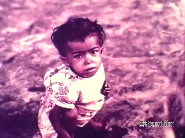 The still from the 1975 CARE World Hunger Fund PSA depicts a young child sitting on the ground, looking directly at the camera. The child appears vulnerable and is wearing a simple outfit with a patterned shirt. The color tones suggest a somber scene, highlighting themes of poverty and hunger. The image evokes empathy and draws attention to the urgent issue of global hunger.
