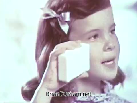 The still features a young girl holding a bar of Ivory soap to her face, likely promoting its qualities. She has wavy hair adorned with a bow and is wearing a patterned dress. The background appears soft and bright, emphasizing the cheerful and clean aesthetic typical of commercials from that era.