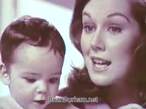 The still features a woman and a baby, likely promoting Ivory Soap. The woman appears engaged and enthusiastic, possibly explaining the benefits of the product. The baby looks intrigued, contributing to the familial and warm atmosphere typical of commercials from that era. The soft, pastel colors enhance the inviting feel of the scene.