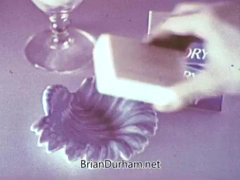 The still from the 1968 Ivory Soap commercial features a hand holding a bar of Ivory soap, poised above a decorative dish. The dish has a shell-like design and is placed on a table, alongside a glass and a box of Ivory Soap in the background, emphasizing the product's branding. The overall color tone appears soft and slightly vintage.