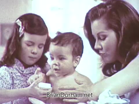 The still from the 1968 Ivory Soap commercial features a woman and two children. The mother is gently holding a baby, while the older girl is applying soap to the baby's skin. The scene conveys a sense of care and hygiene, emphasizing the family-friendly nature of Ivory Soap. The colors are soft and warm, typical of the era’s advertising aesthetic.