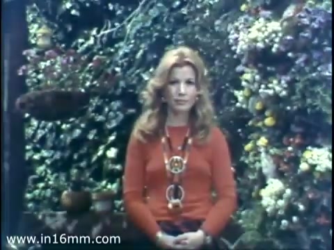 The still features a woman with long, wavy hair seated against a vibrant backdrop of flowers and foliage. She wears an orange sweater adorned with a large necklace. The scene reflects a warm and festive atmosphere, typical of a 1975 Christmas Seal public service announcement.