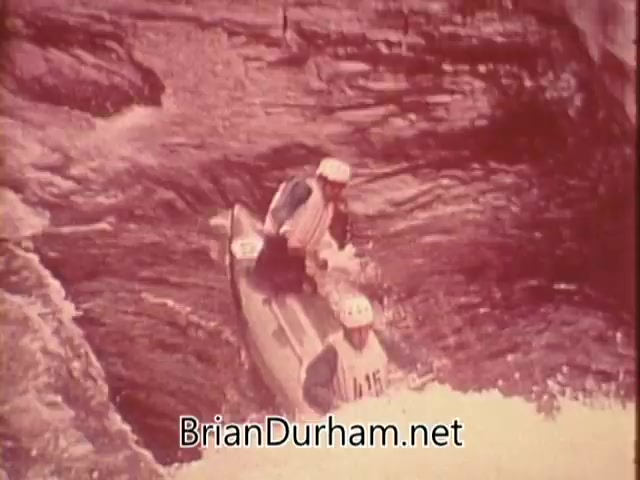 The still from the 1976 US Olympic Committee PSA depicts two individuals in a kayak navigating through turbulent water, possibly in a river setting. They are wearing safety helmets and sports attire, emphasizing the adventurous and competitive spirit of Olympic athletes. The overall tone of the image conveys a sense of determination and teamwork, aligning with the message of supporting the Olympic team by contributing financially. The visual is likely part of a campaign encouraging public involvement and showcasing the dedication of athletes pursuing their dreams.