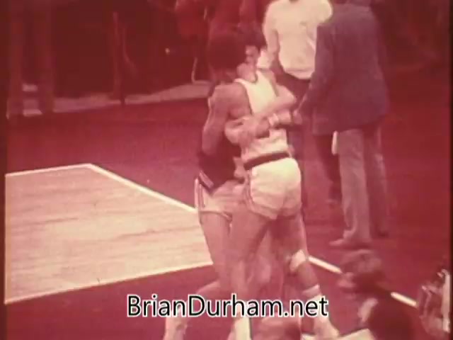 The still from the 'US Olympic Committee PSA - 1976' captures an emotional moment of celebration between athletes on a basketball court. Two players are embracing joyfully, likely after a significant victory. The background hints at a lively sports event, with spectators and officials visible. The PSA emphasizes the sacrifices made by athletes for their Olympic dreams and encourages contributions to support the Olympic team, offering a 'dream of greatness' poster as an incentive for donations.