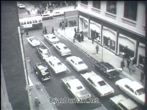 The still from the 1968 'Clean Air PSA' depicts a bustling city street filled with parked and moving cars. The scene captures a mixture of vintage automobiles in various colors, emphasizing the heavy traffic typical of that era. Pedestrians can be seen interacting on the sidewalk, and the overall atmosphere conveys a lively urban environment, likely addressing the growing concerns about air pollution and urban living at the time.