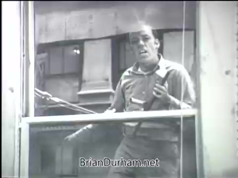 The still from the 'Clean Air PSA - 1968' features a man standing at a window, appearing concerned or distressed. He is dressed in a casual, somewhat disheveled outfit, holding an object in one hand, possibly a tool or piece of equipment. The setting appears urban, with buildings visible outside the window. The overall mood suggests a sense of urgency or a call to action regarding air quality.