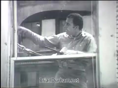 The still from the 'Clean Air PSA - 1968' features a man cleaning a window from inside a building. He appears focused on his task, suggesting a theme of maintaining a clean environment. The monochromatic style emphasizes the message of air quality, typical of public service announcements from that era. The image captures a moment illustrating personal responsibility towards clean living spaces.