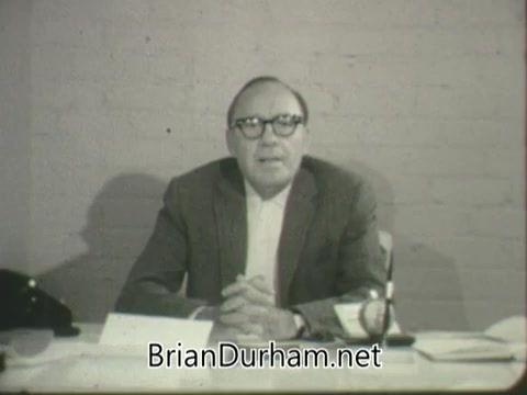 The still features a man seated at a desk, likely Jack Benny, addressing the camera in a straightforward manner. He is wearing glasses and a suit, with a microphone and paperwork visible on the table. The background appears plain, emphasizing his presence. This is a promotional PSA for the Salvation Army from the 1960s, capturing a serious and sincere tone.