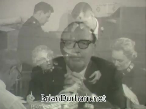 The still from the 'Jack Benny Salvation Army PSA' features a montage of Jack Benny speaking directly to the camera, with a focus on his serious demeanor. In the background, individuals in Salvation Army uniforms are engaged in various activities, possibly supporting community members or organizing efforts. The overall tone seems to convey a message of charity and community support typical of public service announcements from that era.