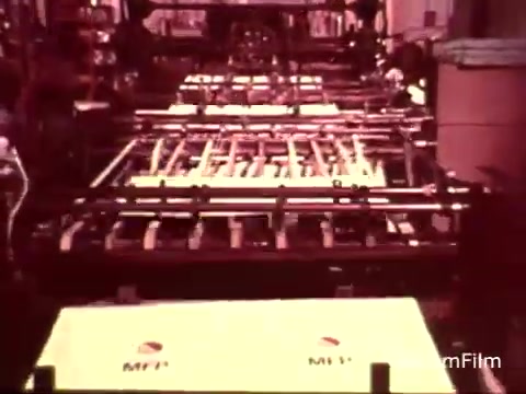 The still features a conveyor system in a factory setting, likely related to the production of Colgate toothpaste with MFP fluoride. It showcases a series of machinery, with boxes labeled 'MFP' moving through the assembly line. The scene captures the industrial process typical of 1970s manufacturing, highlighting the focus on fluoride as a key ingredient in dental care. The color palette and film quality reflect the vintage style of the era.