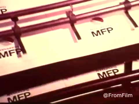 The still features a close-up of a production line or packaging area, prominently displaying boxes labeled 'MFP.' The scene reflects the vintage aesthetic of a 1970s Colgate toothpaste commercial, emphasizing the incorporation of MFP fluoride in their product. The warm color tones typical of that era contribute to its nostalgic feel. The image showcases the brand's focus on dental health and innovation.