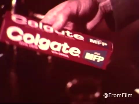 The still features a close-up of a person's hand holding a tube of Colgate toothpaste prominently labeled 'MFP.' The tube has a bright red color with bold white lettering. This commercial, from the 1970s, emphasizes the use of MFP fluoride, suggesting a focus on dental health and protection. The vintage film quality and color saturation evoke a nostalgic feel typical of that era's advertisements.
