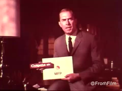 The still features a man in a suit, holding a sign that promotes Colgate toothpaste with MFP fluoride, indicative of a 1970s advertising style. The setting appears to be a living room, with a lamp in the background contributing to the vintage aesthetic. The focus is on the product being endorsed, emphasizing its fluoride content.