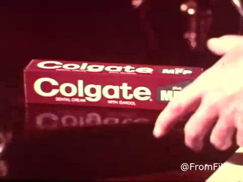 The still from the 1970s Colgate toothpaste commercial displays a red Colgate toothpaste box prominently featuring the 'MFP' designation. A hand is seen pointing toward the box, highlighting the product. The vintage aesthetic of the 16mm print adds to the nostalgic feel, evoking a classic advertising style from that era. The focus is on promoting the dental cream with an emphasis on fluoride.