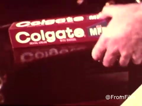 The still features a close-up shot of a hand reaching for a box of Colgate toothpaste, specifically highlighting the MFP fluoride variant. The toothpaste packaging is prominently displayed, showcasing its retro design typical of the 1970s. The overall aesthetic reflects the era's advertising style, emphasizing dental health and fluoride benefits.