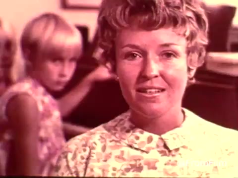 The still from the 1970s Colgate toothpaste commercial showcases a woman with a friendly demeanor in a floral-patterned blouse, smiling as she speaks. Behind her, a young girl with blonde hair and a serious expression appears, possibly engaged in a piano activity. The overall aesthetic reflects the era's style, with warm colors and a domestic setting, emphasizing family and dental care. This particular advertisement likely promotes the benefits of MFP fluoride in Colgate toothpaste.