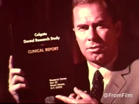 The still appears to depict a man in formal attire holding a black report titled 'Colgate Dental Research Study CLINICAL REPORT.' The setting suggests a promotional or informational tone typical of 1970s advertisements, likely emphasizing the benefits of Colgate toothpaste with MFP fluoride. The image captures a moment geared towards instilling confidence in the product through research findings.