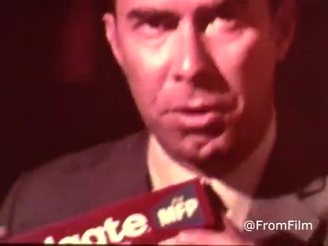 The still from the 'Colgate Toothpaste Commercial - with MFP fluoride! 1970's' features a man in a suit, closely holding a tube of Colgate toothpaste. The lighting creates a warm, slightly dramatic atmosphere. He appears to be emphasizing the product, likely promoting its benefits. The image reflects typical commercial aesthetics of the 1970s.