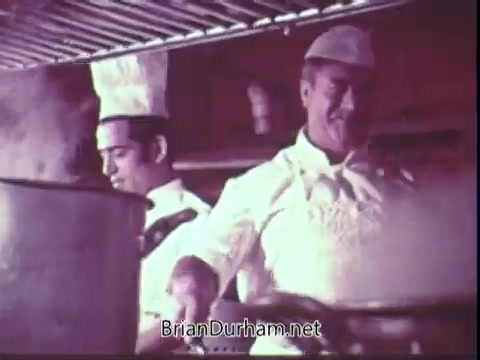 The still from the 1972 Blue Bonnet Margarine commercial features two chefs in a kitchen setting. One chef is focused on stirring a large pot, while the other, wearing a white chef's hat and uniform, appears to be observing or assisting. The kitchen is steamy, suggesting cooking activity, which highlights the use of Blue Bonnet Margarine in culinary preparations. The overall atmosphere conveys a sense of professionalism and culinary expertise.