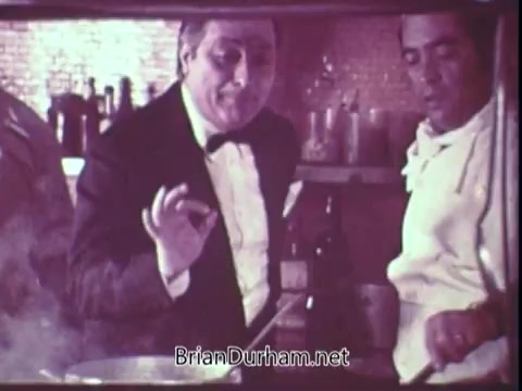 The still from the 1972 Blue Bonnet Margarine commercial features two men in a kitchen setting. One is wearing a tuxedo and confidently gestures while talking, possibly as part of a cooking demonstration. The other, dressed in a chef’s attire, appears to be focused on a pot or pan, suggesting a collaborative cooking scene. The atmosphere is warm and inviting, with elements typical of a vintage kitchen, and hints of steam rising from the cooking surface.